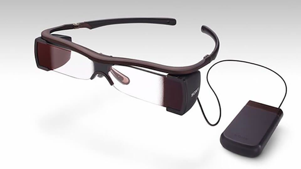 Em uma superfície cinza, óculos que exibe legendas na lente, ao lado aparelho para recepção de audiodescrição